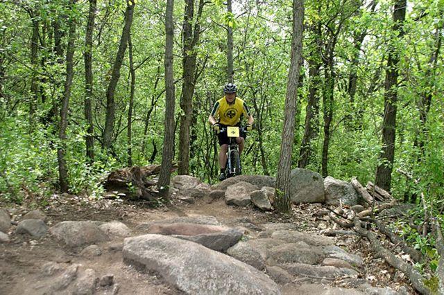 A mountain biker navigating a rocky trail in a dense forest, surrounded by green trees and foliage. The biker is wearing a yellow and black jersey and helmet, focused on the rocky terrain ahead. Lebanon Hills mountain bike trail.