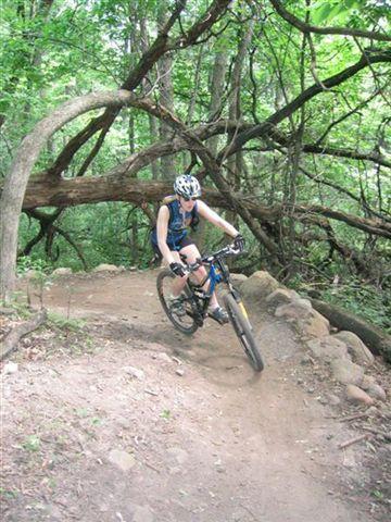 A person riding a mountain bike on a winding dirt trail surrounded by lush green foliage and overhanging trees. The rider is wearing a helmet and is leaning into a turn as they navigate the rugged terrain. Lebanon Hills mountain bike trail.