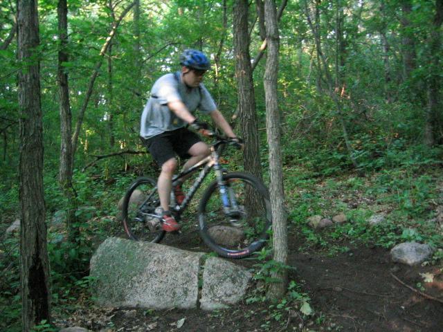 A person riding a mountain bike over a large rock on a forest trail, surrounded by trees and greenery. The rider is wearing a blue helmet and casual athletic clothing. Lebanon Hills mountain bike trail.