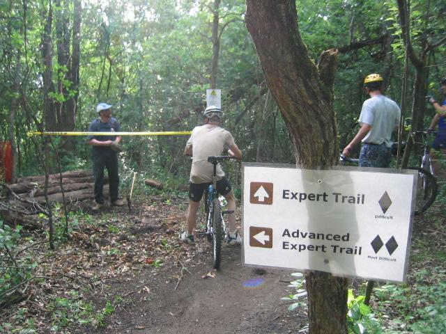 A group of mountain bikers at a trail intersection in a wooded area, with a sign indicating directions for the "Expert Trail" and "Advanced Expert Trail." One biker is stopped on their bike, while two others are discussing near a yellow caution tape stretched across the path. The environment is lush with trees and underbrush. Lebanon Hills mountain bike trail.