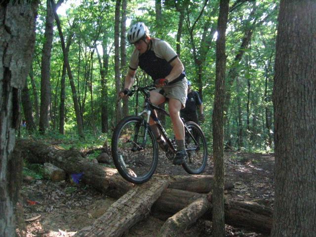 A cyclist in a helmet and athletic wear is airborne, jumping over a log on a mountain bike trail surrounded by trees in a lush forest. Sunlight filters through the leaves, illuminating the scene. Lebanon Hills mountain bike trail.