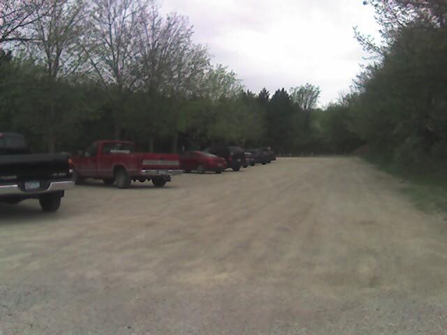 A gravel parking area lined with several parked pickup trucks and cars, surrounded by green trees under a cloudy sky. Lebanon Hills mountain bike trail.