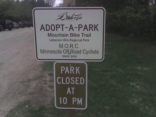 Sign at Lebanon Hills Regional Park indicating the "Adopt-A-Park" mountain bike trail, managed by the Minnesota Off-Road Cyclists (M.O.R.C) since 2005. A second sign below notes that the park closes at 10 PM. The background features grassy areas and a parking lot. Lebanon Hills mountain bike trail.