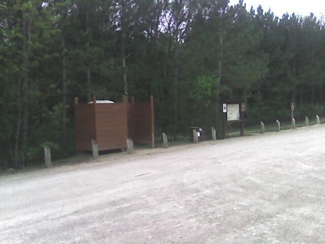 Image of a wooded area with a gravel road. In the foreground, there is a small wooden structure, likely a restroom, situated beside a sign post. The background features dense trees, indicating a natural setting, with additional informational signs visible nearby. Lebanon Hills mountain bike trail.