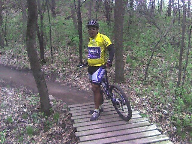 A cyclist in a bright yellow jersey stands on a wooden bridge in a forested area, surrounded by green trees and foliage. The cyclist is wearing a helmet and sunglasses, with a mountain bike beside them. A dirt path is visible in the background, indicating a trail for biking. Lebanon Hills mountain bike trail.