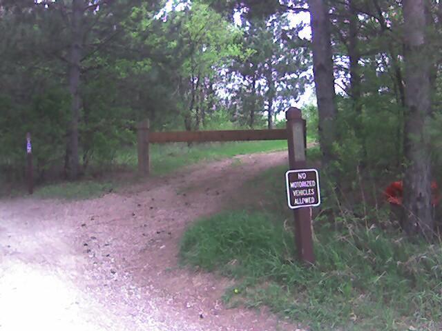Gravel path leading into a wooded area, with a wooden barrier across the entrance and a sign stating "No Motorized Vehicles Allowed" on the right. Lush greenery surrounds the entrance, indicating a natural setting. Lebanon Hills mountain bike trail.