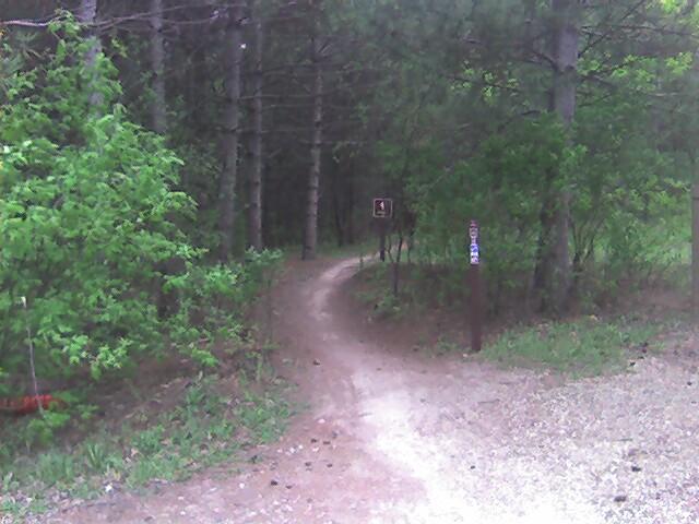 A winding dirt pathway leading into a forested area, surrounded by greenery and trees. A signpost is visible along the path, partially obscured by foliage. The scene depicts a tranquil outdoor setting, inviting exploration. Lebanon Hills mountain bike trail.