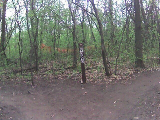 A dirt trail fork in a wooded area, with a sign indicating directions. The surrounding trees are lush and green, and some orange fencing is visible in the background. Lebanon Hills mountain bike trail.