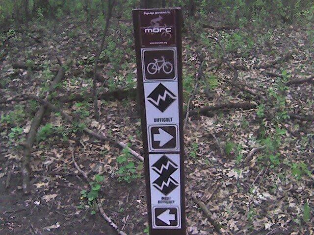 A trail sign in a wooded area featuring symbols indicating biking accessibility and difficulty levels. The sign includes icons for bicycle use, different difficulty ratings (easy, moderate, and most difficult), and arrows showing directional guidance. Surrounding the sign are trees and foliage typical of a forest environment. Lebanon Hills mountain bike trail.