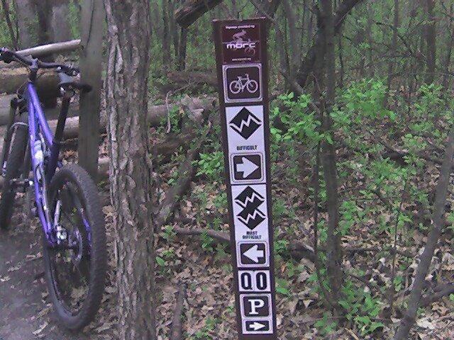 A trail sign in a wooded area, featuring symbols indicating bike usage, difficulty levels, and directional arrows. A purple mountain bike is leaned against a tree nearby. Lebanon Hills mountain bike trail.