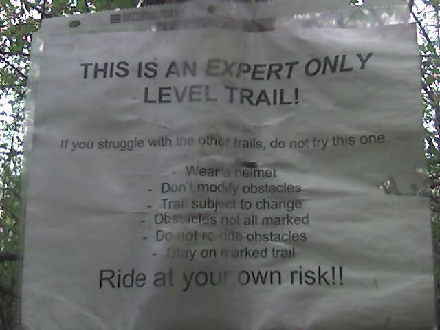 A worn sign warning cyclists that the trail is for expert riders only. The sign advises wearing a helmet, not modifying obstacles, and indicates that the trail may change and not all obstacles are marked. It warns users to ride at their own risk. Lebanon Hills mountain bike trail.