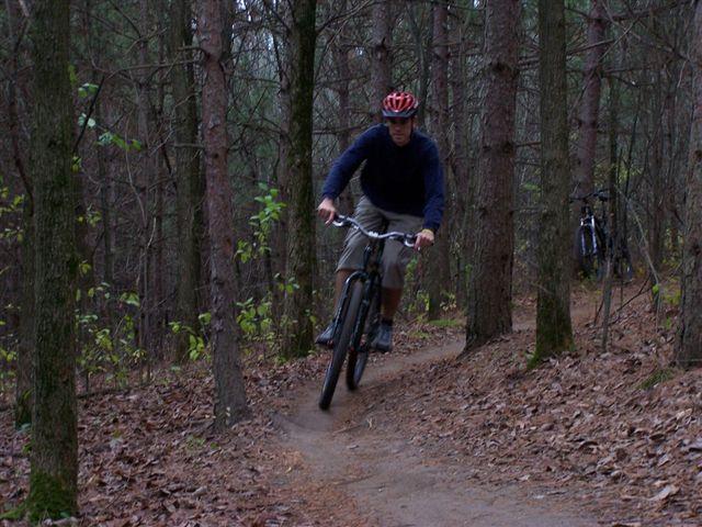A cyclist riding a mountain bike on a dirt trail surrounded by tall trees in a wooded area, with another bike parked in the background. The rider is wearing a red helmet and is focused on navigating the trail. Lebanon Hills mountain bike trail.