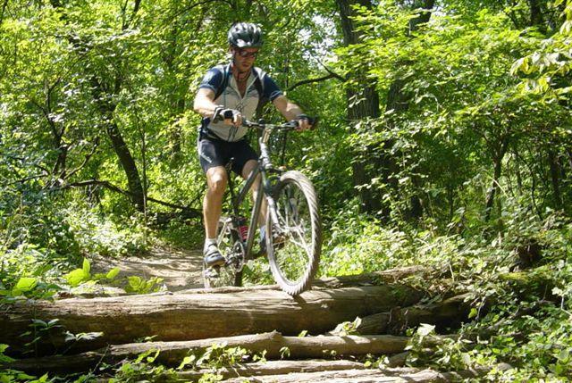 A mountain biker navigating over logs on a forest trail, surrounded by lush green vegetation. The cyclist is wearing a helmet and athletic gear while riding a mountain bike. Lebanon Hills mountain bike trail.