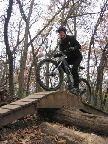 A mountain biker navigating a wooden bridge in a forested area, surrounded by trees with autumn foliage. The rider is wearing a helmet and dark clothing, showcasing skill as they balance on the bridge. Lebanon Hills mountain bike trail.