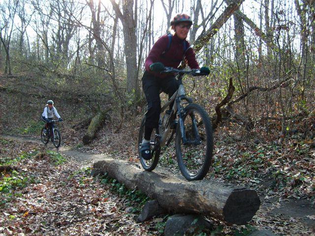 Two mountain bikers navigating a wooded trail. One biker is confidently balancing on a log, while the second biker follows behind on the path. The setting is surrounded by trees, with fallen leaves on the ground, suggesting a natural outdoor environment. Lebanon Hills mountain bike trail.