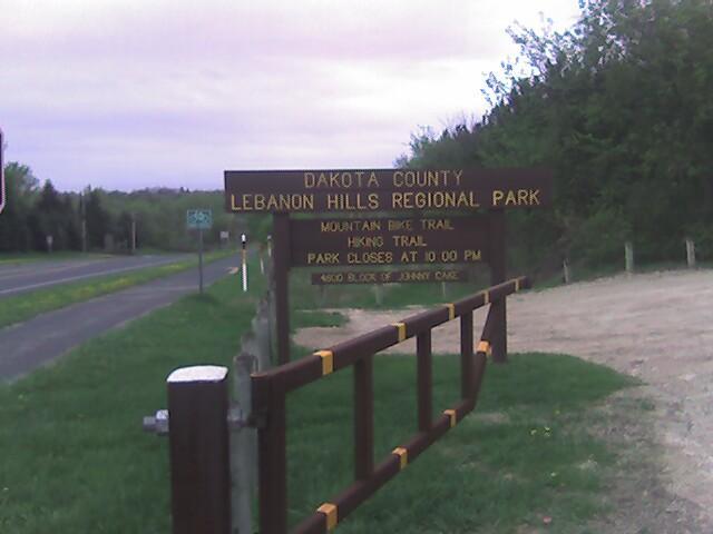 Sign marking the entrance to Dakota County's Lebanon Hills Regional Park, featuring information about mountain biking and hiking trails, as well as the park's closing time of 10:00 PM. The sign is positioned near a road, surrounded by greenery. Lebanon Hills mountain bike trail.