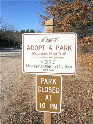 A sign for the Adopt-A-Park Mountain Bike Trail located in Lebanon Hills Regional Park, indicating that the park is closed at 10 PM. The sign also mentions the Minnesota Off-Road Cyclists (M.O.R.C.) partnership since 2005. The background includes trees and a gravel path. Lebanon Hills mountain bike trail.