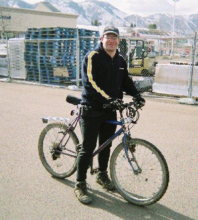 A person wearing a dark jacket with a yellow stripe stands beside a purple mountain bike in an outdoor setting, with industrial structures and mountains in the background. Red Rocks / Dakota Ridge mountain bike trail.