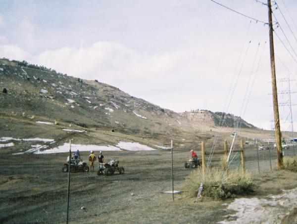 A group of four all-terrain vehicles (ATVs) on a dirt track, with a mountainous landscape in the background. Some snow patches are visible on the ground, and several power lines run alongside the track. The sky is overcast, indicating a cloudy day. Red Rocks / Dakota Ridge mountain bike trail.