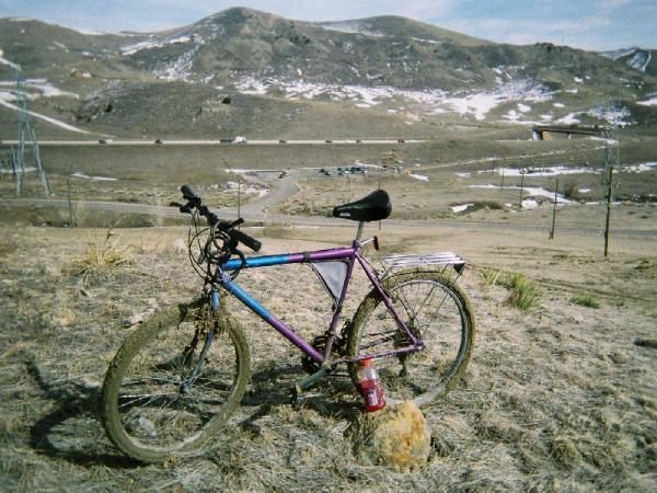 A colorful mountain bike with a purple and blue frame, leaning against a rock in an open grassy area. In the background, rolling hills with patches of snow and a winding dirt road can be seen. The sky is partly cloudy, indicating a cool outdoor setting. Red Rocks / Dakota Ridge mountain bike trail.