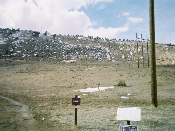 A barren landscape with a gentle incline leading to a snowy hillside. In the foreground, there is a brown sign labeled "Zorro" on the left side of the image. To the right, a white sign is visible beside a dirt path, and utility poles line the background against a cloudy blue sky. Red Rocks / Dakota Ridge mountain bike trail.