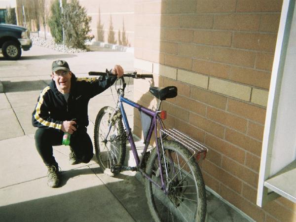 A smiling person in casual attire, including a black jacket with yellow stripes and a baseball cap, kneels beside a weathered purple bicycle parked against a brick wall. The surrounding area appears to be an outdoor space, and a pickup truck is visible in the background. Red Rocks / Dakota Ridge mountain bike trail.