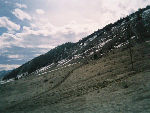 A mountainous landscape featuring a sloping hillside with patches of snow, sparse vegetation, and a partly cloudy sky. A dirt path winds through the foreground, leading up the slope, while a power pole stands to the right. Red Rocks / Dakota Ridge mountain bike trail.