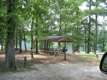 A wooden pavilion surrounded by trees at a park, with picnic tables underneath and a few people in the area. There is a gravel path leading to the pavilion, and a calm body of water is visible in the background. Oak Mountain State Park Bump Trail mountain bike trail.