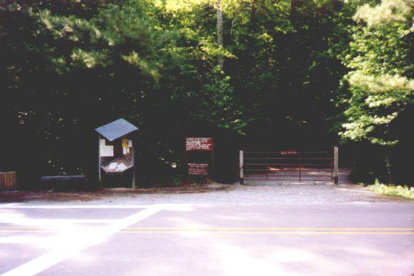 A gravel road entrance to a forested area, featuring a wooden gate on the right and an information board on the left with various notices and maps. The surroundings are rich with green trees, indicating a natural setting. Oak Mountain State Park mountain bike trail.