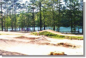 A sunny outdoor scene featuring a dirt bike track with jumps and hills, surrounded by tall trees. In the background, a group of people can be seen near a lake, creating a peaceful, recreational atmosphere. Oak Mountain State Park Bump Trail mountain bike trail.