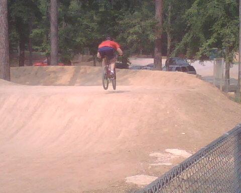 A person riding a bicycle is airborne over a dirt ramp in a bike park, surrounded by trees and a fence. Oak Mountain State Park mountain bike trail.