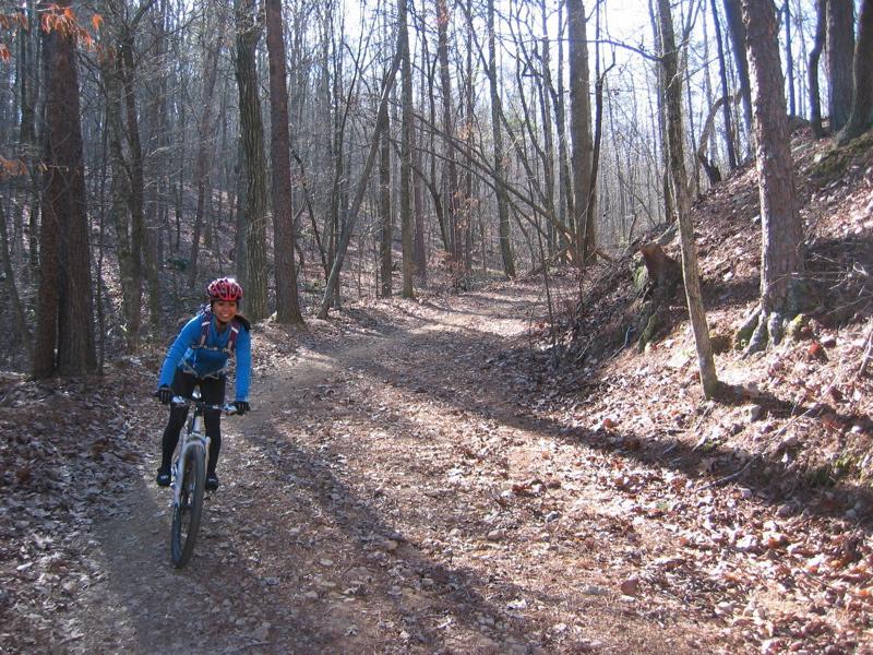 A young person riding a mountain bike on a dirt trail surrounded by bare trees in a wooded area, with fallen leaves on the ground and sunlight filtering through the branches. Oak Mountain State Park mountain bike trail.