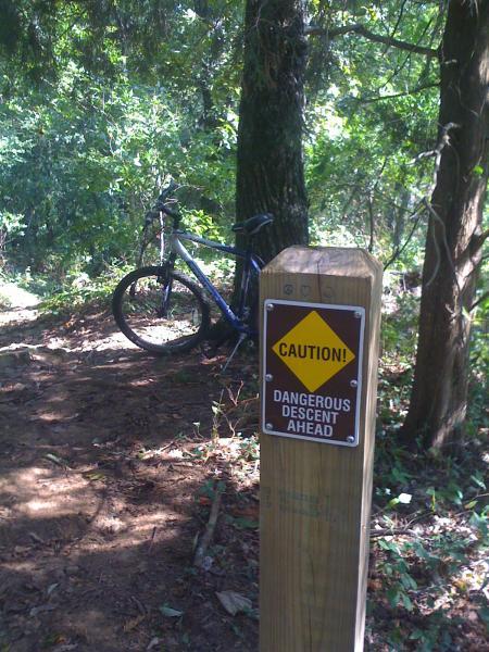 A mountain bike parked next to a wooden signpost in a wooded area, displaying a caution sign that reads "CAUTION! DANGEROUS DESCENT AHEAD." The surroundings are lush with trees and vegetation, indicating a trail or path through the forest. Monte Sano State Park &amp; Land Trust mountain bike trail.