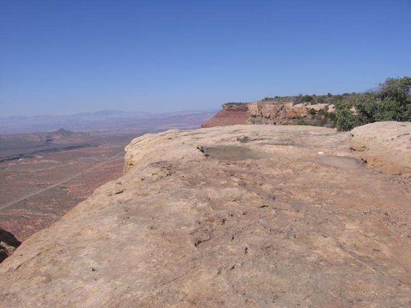 A panoramic view from a rocky cliff edge overlooking a vast desert landscape, with clear blue skies and distant mountains on the horizon. Gooseberry Mesa mountain bike trail.