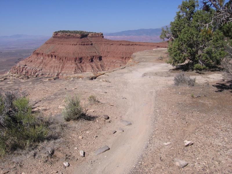A rocky trail leading towards a prominent red rock formation, with sparse desert vegetation and a clear blue sky in the background. The formation features layered red cliffs and a flat top, surrounded by a vast, arid landscape. Gooseberry Mesa mountain bike trail.