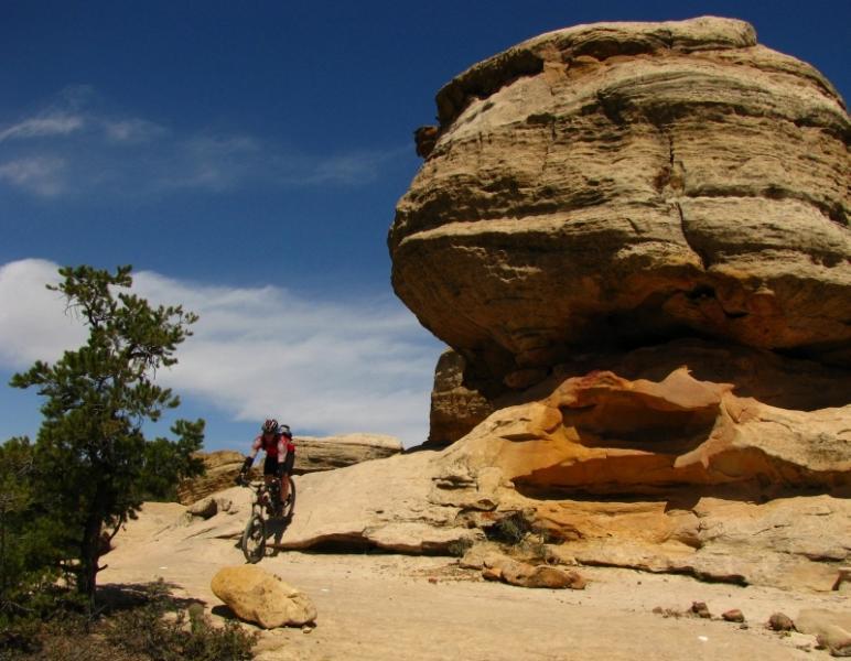 A mountain biker navigating a rocky terrain near a large, rounded rock formation, with blue skies and scattered clouds in the background. Gooseberry Mesa mountain bike trail.