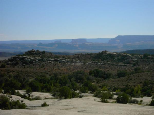 A panoramic view of a rocky landscape featuring a mix of low shrubs and larger boulders in the foreground, with distant cliffs and a blue sky in the background. The scene conveys a sense of rugged natural beauty. Klondike Bluffs mountain bike trail.