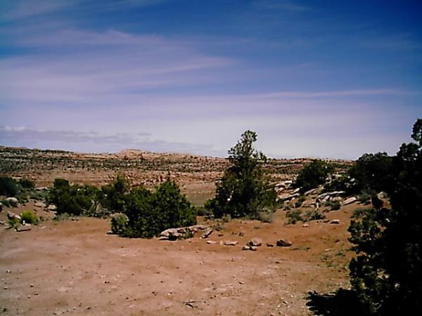 A scenic view of a dry, desert landscape featuring rocky terrain, sparse vegetation, and a clear blue sky with wispy clouds. The foreground includes scattered shrubs and boulders, leading to distant hills and mesas in the background. Klondike Bluffs mountain bike trail.