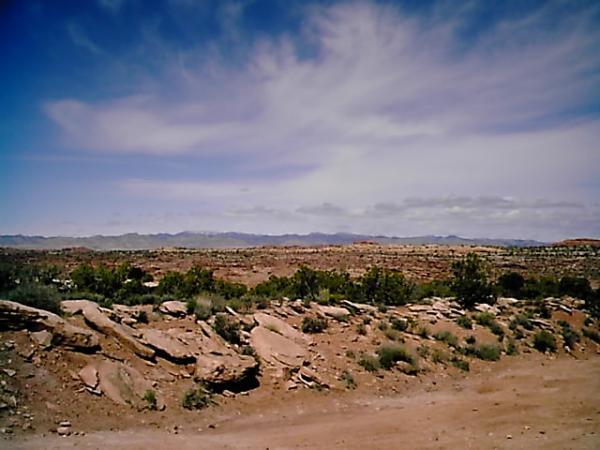 A scenic view of a rocky landscape under a partly cloudy sky, featuring desert vegetation and distant mountains on the horizon. The foreground includes various sized rocks and sparse greenery, with a dirt path winding through the terrain. Klondike Bluffs mountain bike trail.