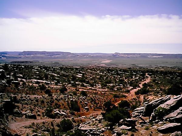 A panoramic view of a desert landscape featuring rocky terrain, sparse vegetation, and distant mesas under a clear blue sky. The scene captures the natural beauty of the arid environment, with winding paths and scattered shrubs throughout the foreground. Klondike Bluffs mountain bike trail.