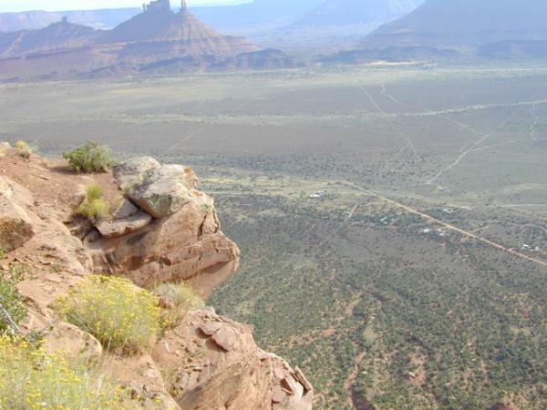A panoramic view from a rocky cliff overlooking a vast, arid landscape. The foreground features a rugged rock formation with patches of yellow wildflowers, while the background showcases distant mesas and plateaus under a clear blue sky. The expansive terrain includes winding dirt roads and sparse vegetation stretching to the horizon. Porcupine Rim mountain bike trail.