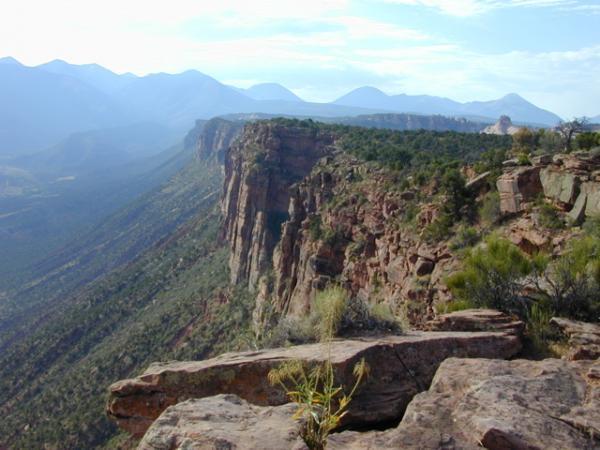A panoramic view of a rugged cliffside with a rocky outcrop in the foreground, surrounded by lush greenery and distant mountains under a partly cloudy sky. The landscape showcases the natural beauty of the terrain, emphasizing the steep cliffs and rolling hills. Porcupine Rim mountain bike trail.