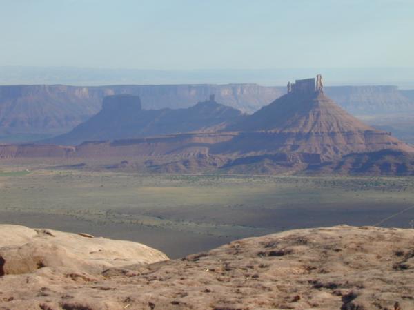A panoramic view of rugged desert landscape featuring mesas and cliffs under a clear blue sky. The foreground shows rocky terrain leading to distant mesas with steep sides and flat tops, creating a dramatic backdrop. The scene captures the tranquility and vastness of the natural environment. Porcupine Rim mountain bike trail.