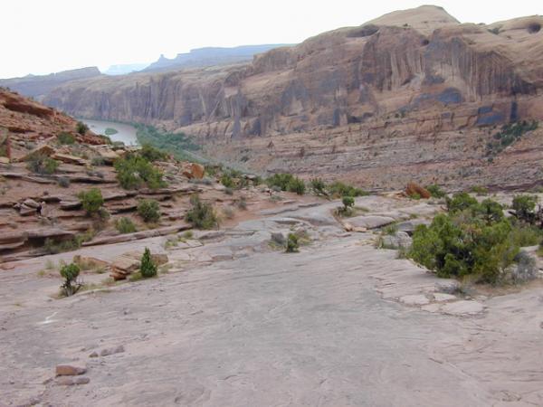 A rocky landscape with layered cliffs and a winding river below. Sparse vegetation, including small bushes, is visible among the rocks, and the sky is overcast with distant mountains in the background. The scene captures a rugged, natural terrain typical of a desert or canyon environment. Porcupine Rim mountain bike trail.