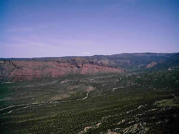 A vast desert landscape with rolling hills and colorful rock formations under a clear blue sky. The foreground features arid terrain with sparse vegetation, while the background showcases layered cliffs in shades of red and purple, extending into the distance. Porcupine Rim mountain bike trail.