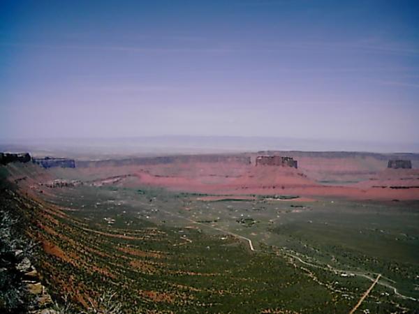 A panoramic view of a vast desert landscape featuring red rock formations and rolling hills under a clear blue sky. The foreground showcases green vegetation, while the distant mesas and cliffs create a striking horizon. Porcupine Rim mountain bike trail.