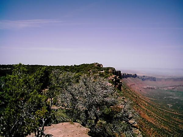 A panoramic view of a rugged landscape featuring a rocky cliff edge covered with sparse vegetation, overlooking a vast expanse of rolling hills and distant mountains under a clear blue sky. Porcupine Rim mountain bike trail.