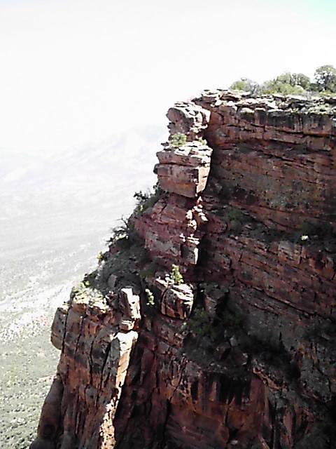 A rugged rock formation juts out from a steep cliff, showcasing layers of reddish-brown stone. Sparse vegetation clings to the edges, and a vast mountainous landscape stretches into the distance under a clear sky. Porcupine Rim mountain bike trail.