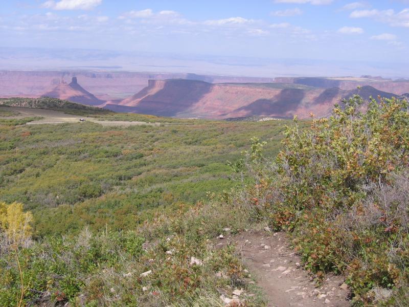 A panoramic view of a rugged landscape featuring red rock formations and a vast, open sky. In the foreground, a grassy area with bushes and a dirt path leads towards the distant cliffs, showcasing a combination of greenery and rocky terrain under a partly cloudy sky. Porcupine Rim mountain bike trail.