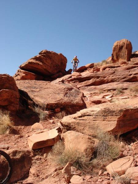 A mountain biker navigating a rocky terrain under a clear blue sky, with large red rock formations and sparse vegetation in the background. Porcupine Rim mountain bike trail.